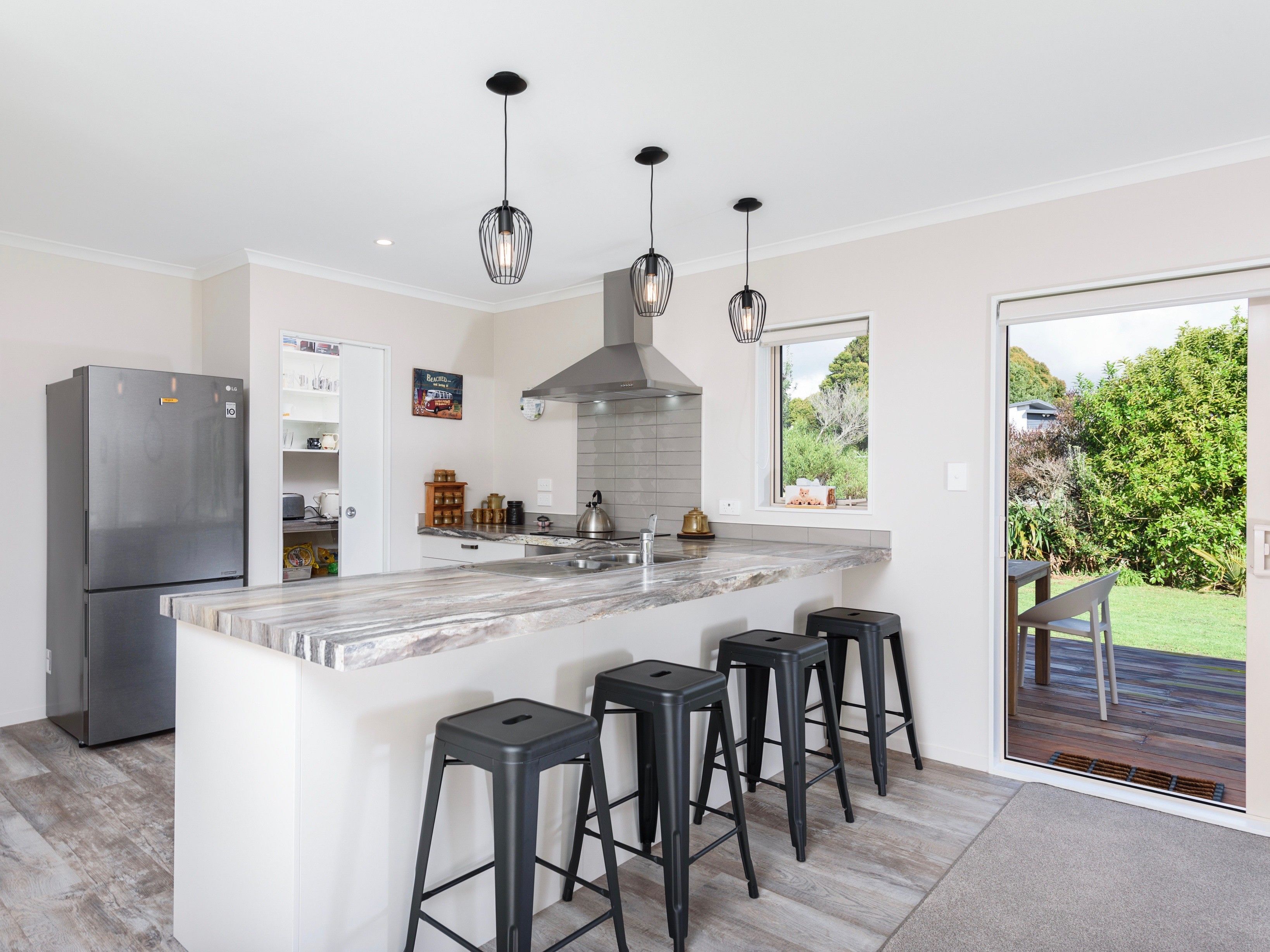 A modern kitchen with a central island, black bar stools, a stainless steel refrigerator, and a view of the garden through a sliding door.