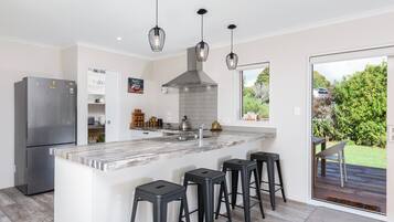 A modern kitchen with a central island, black bar stools, a stainless steel refrigerator, and a view of the garden through a sliding door.