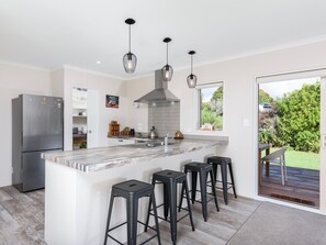 A modern kitchen with a central island, black bar stools, a stainless steel refrigerator, and a view of the garden through a sliding door.