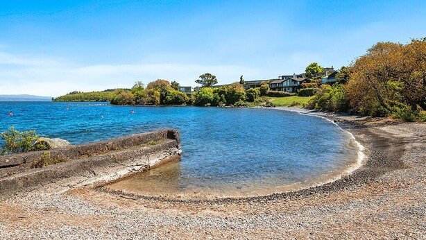 Un paysage côtier avec une plage de galets, un ciel bleu dégagé et un bâtiment doté d’un jardin et d’arbres en arrière-plan.