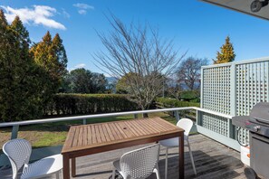 A wooden deck with a table and chairs, a grill, and a view of a landscaped garden and trees. - Te Kopua - Acacia Bay Holiday Home (Acacia Bay)