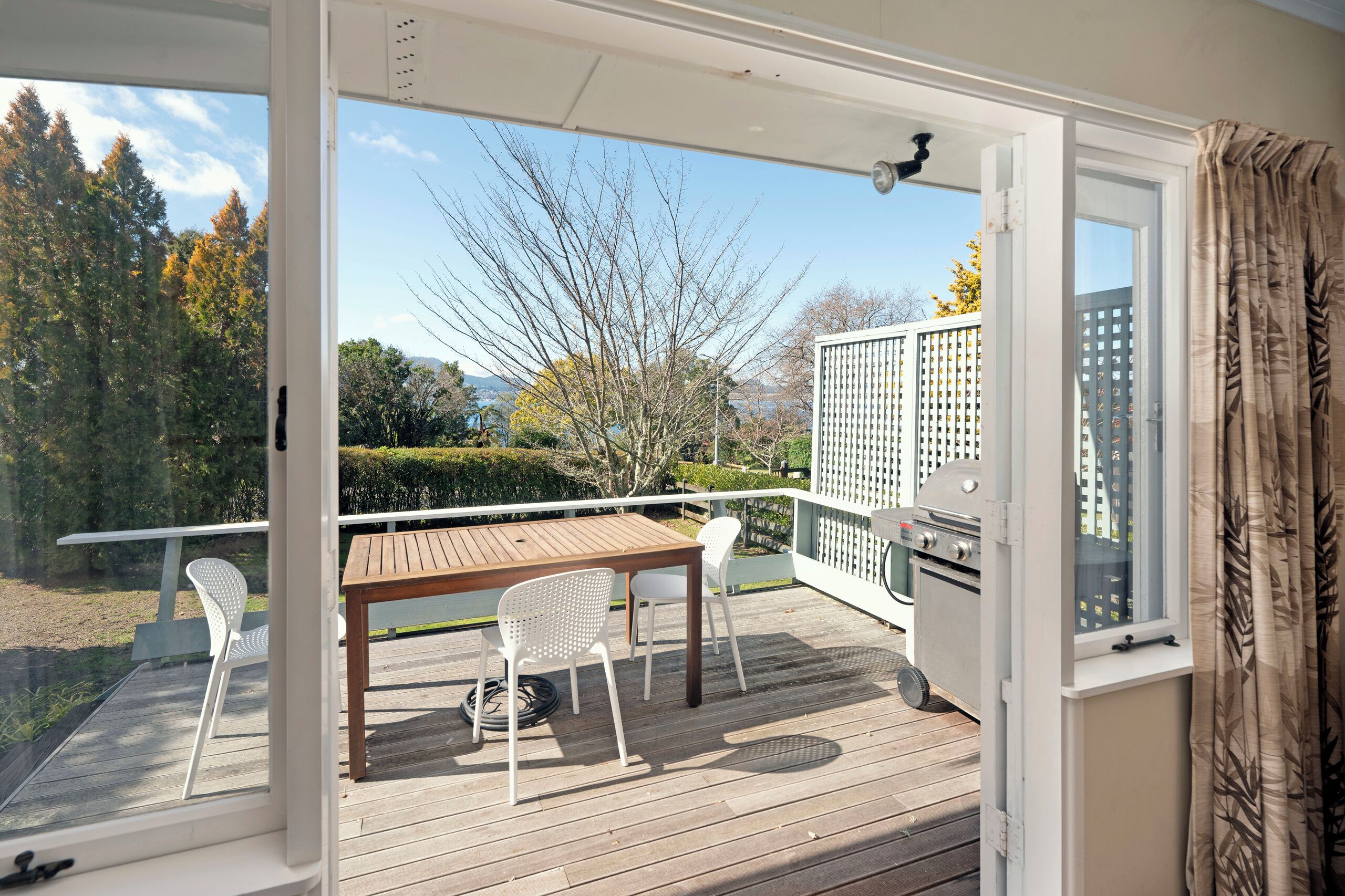 Una terraza de madera con mesa y sillas, una parrilla y vistas a árboles y edificios.