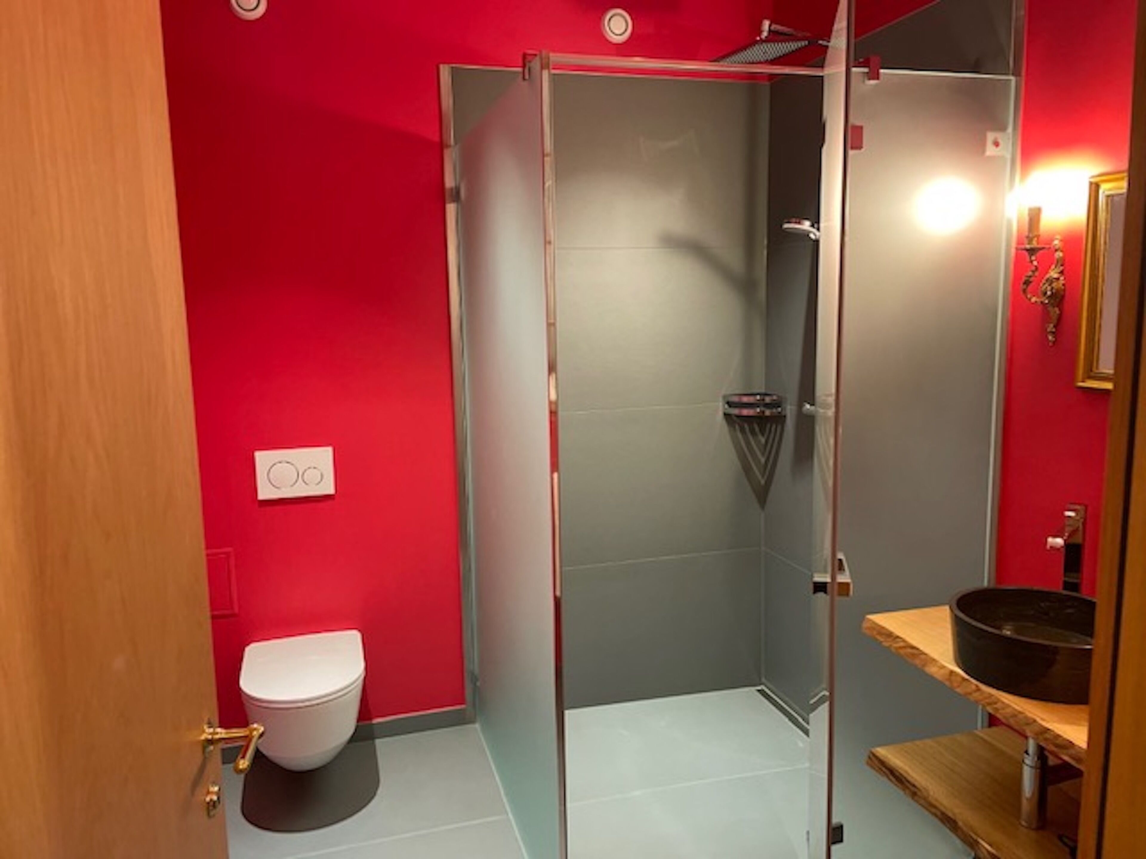 Ein Badezimmer mit roter Wand, einer weißen Toilette, einer Duschkabine aus Glas und einem hölzernen Waschtisch mit schwarzem Becken.