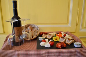 A table with a bottle of wine, two wine glasses, a basket of bread, and a variety of cheeses and vegetables.