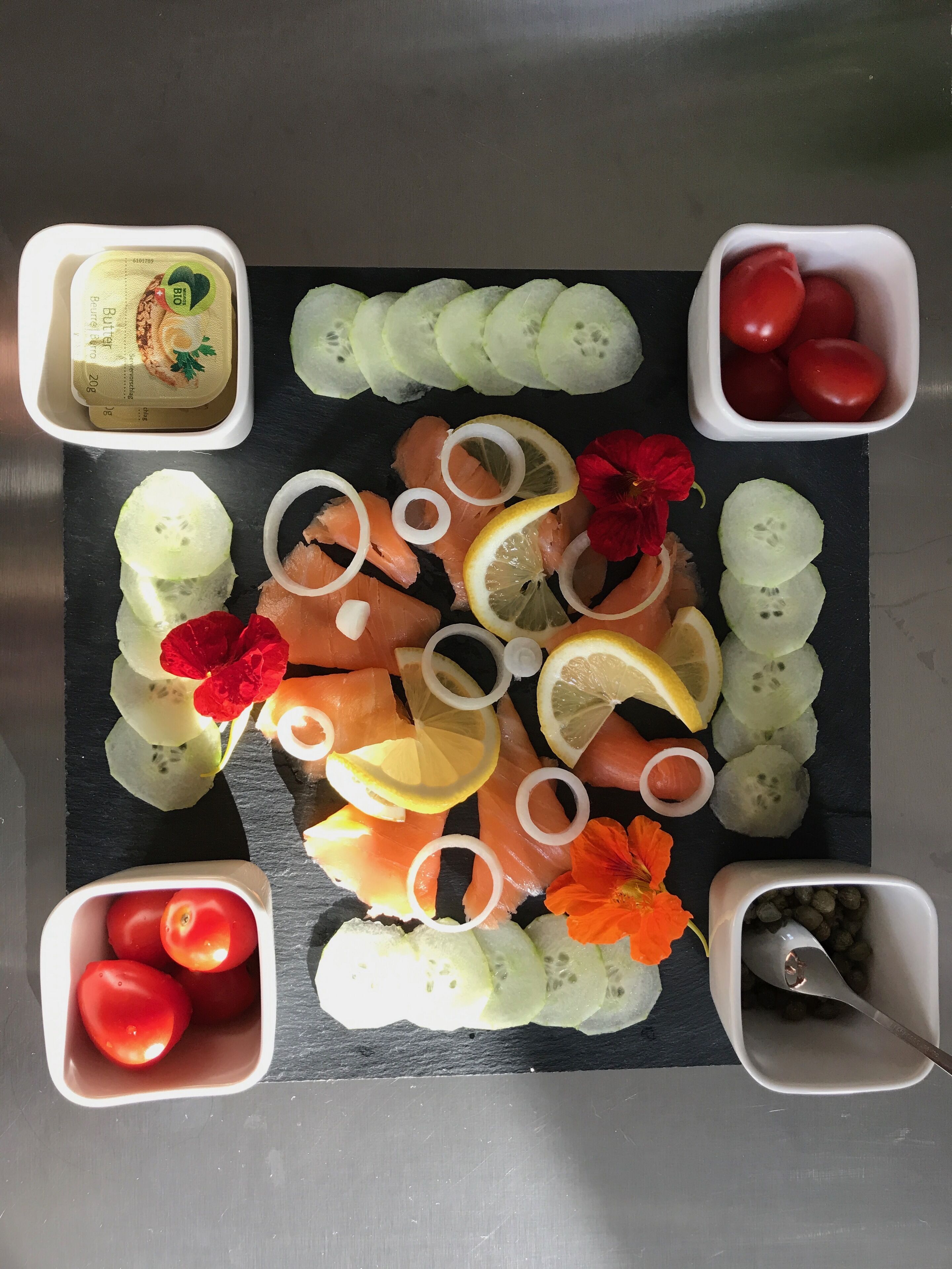 Una bandeja con salmón ahumado, rodajas de pepino, gajos de limón y tomates cherry dispuestos sobre una tabla de piedra negra.
