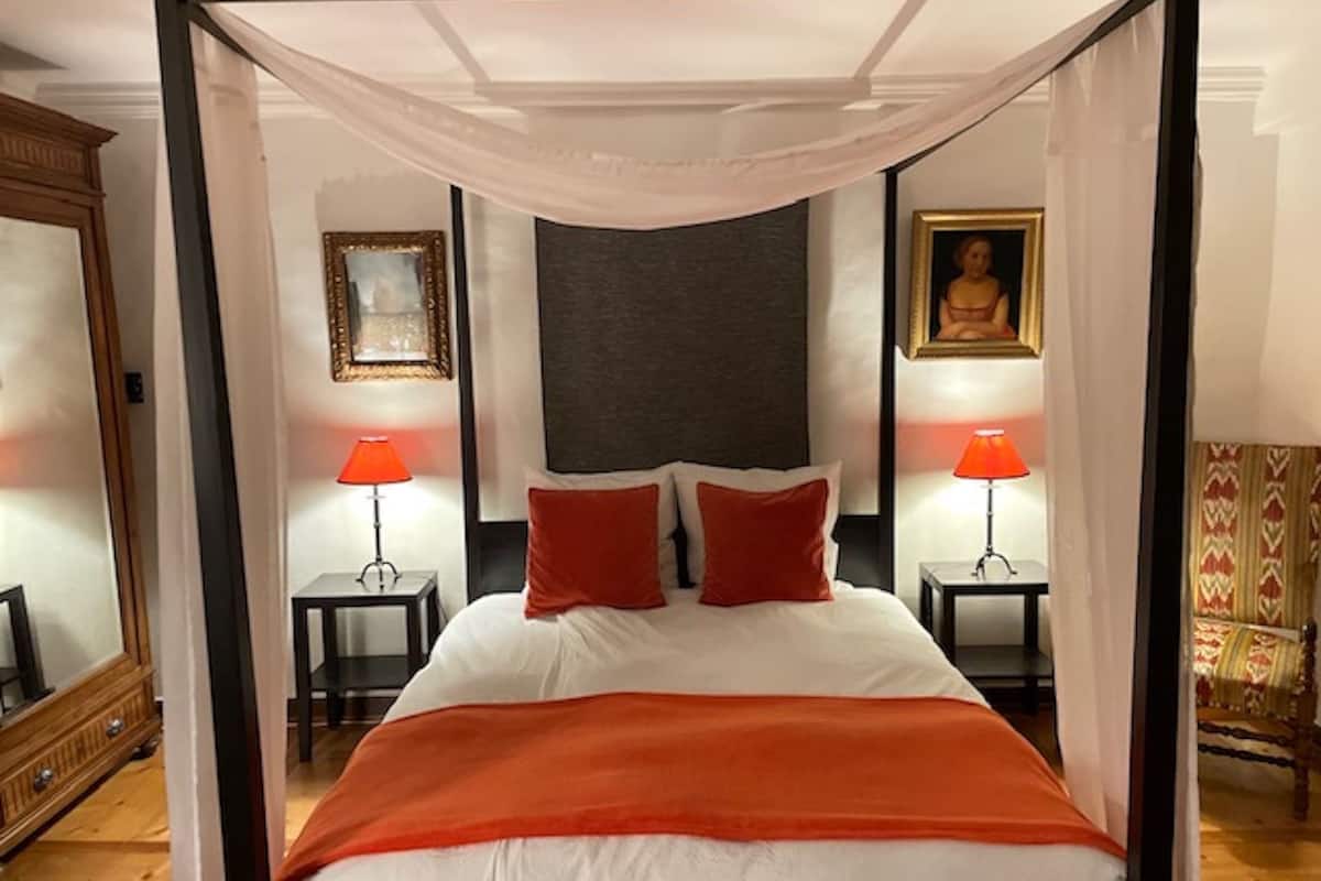 Ein Bett mit Baldachin, weißes Blätterdach, zwei rote Kissen und eine rote Tagesdecke. Zwei Nachttische mit Lampen, ein gerahmtes Bild an der Wand und ein Spiegel links.