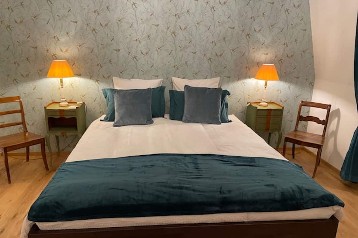 Ein ordentlich bezogenes Bett mit einem petrolfarbenen Überwurf, zwei Nachttische mit Lampen und einem blumigen Wandpapier.
