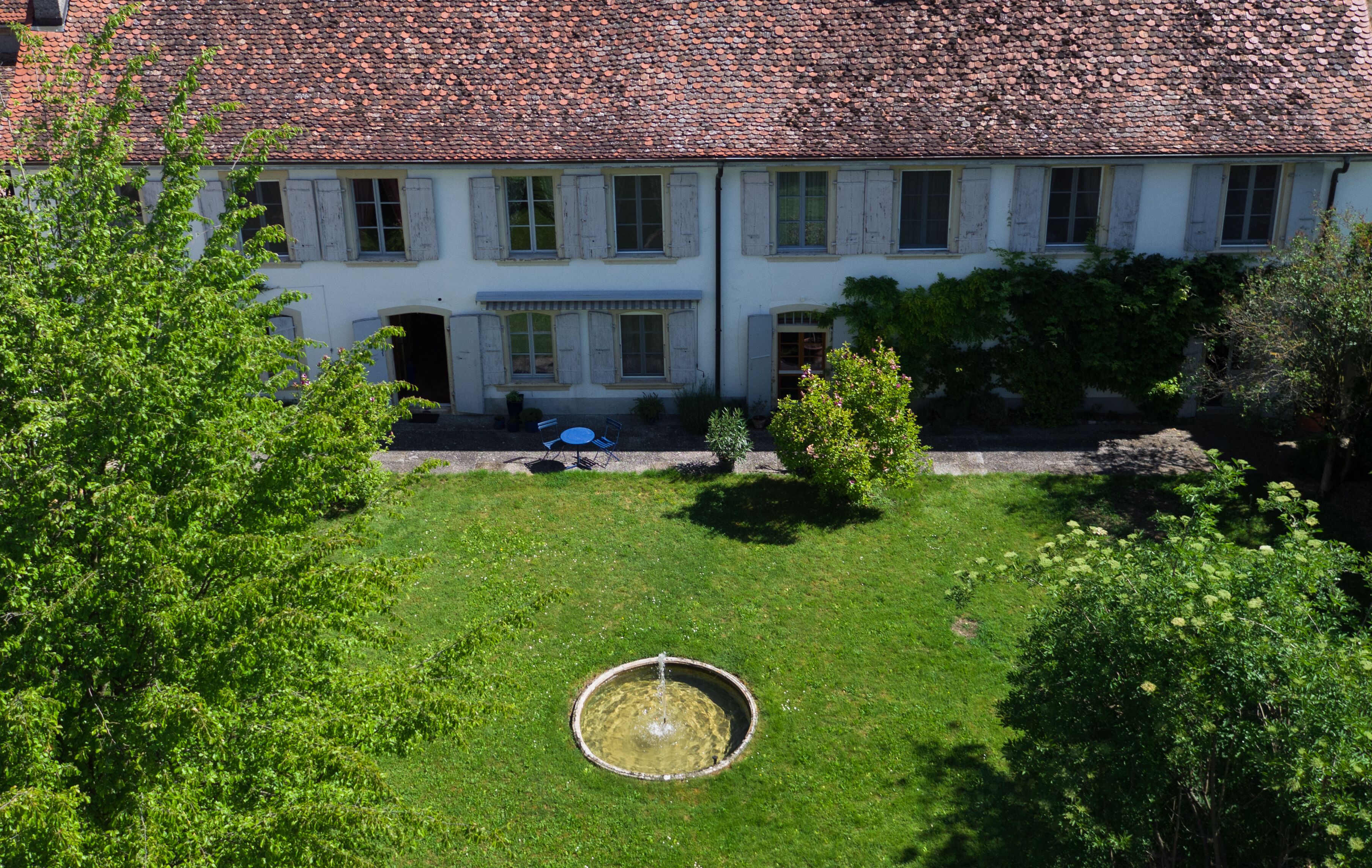 A large, white building with a tiled roof and a circular fountain in the center of a well-maintained lawn.