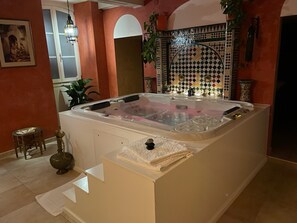Ein Wellnessbereich mit einer großen Badewanne, einem kleinen Tisch mit einer Flasche und einer Wand mit gemusterten Fliesen.