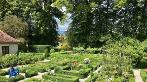 Un jardín meticulosamente cuidado con setos geométricos y plantas en flor, un pequeño edificio y una vista despejada del cielo.