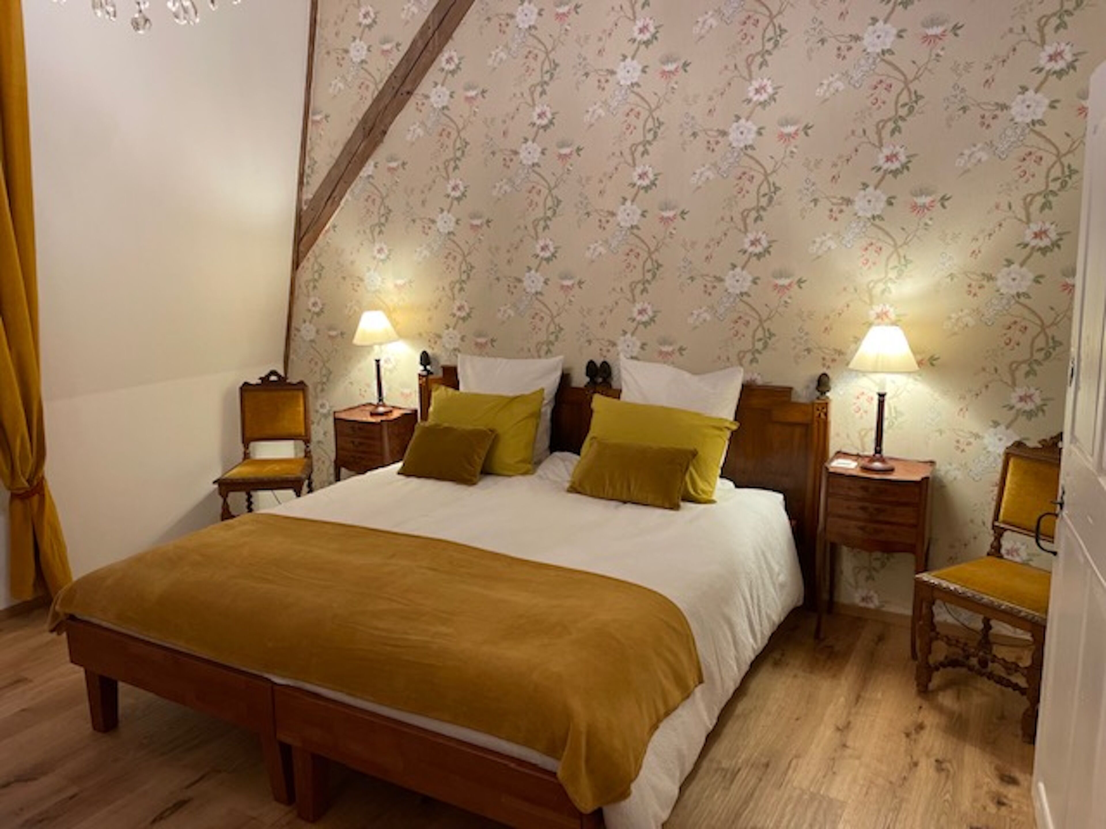 Une chambre avec un grand lit, deux tables de chevet avec des lampes, un banc en bois, un papier peint à motifs floraux et une poutre en bois au plafond.
