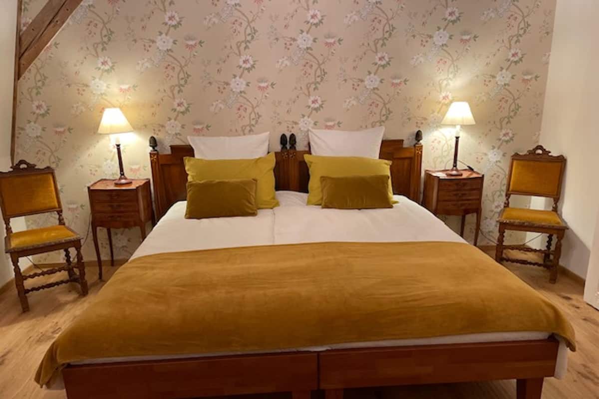 Ein Doppelbett mit einer senfgelben Decke, zwei Nachttische mit Lampen, blumiger Wandtapete und zwei Holzstühle.