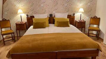A double bed with a mustard-colored blanket, two bedside tables with lamps, floral wallpaper, and two wooden chairs.