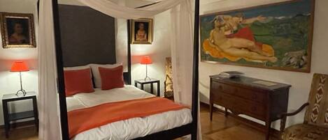 A four-poster bed with white and orange bedding, two bedside tables with lamps, a wooden desk, a chair, and a large landscape painting on the wall.