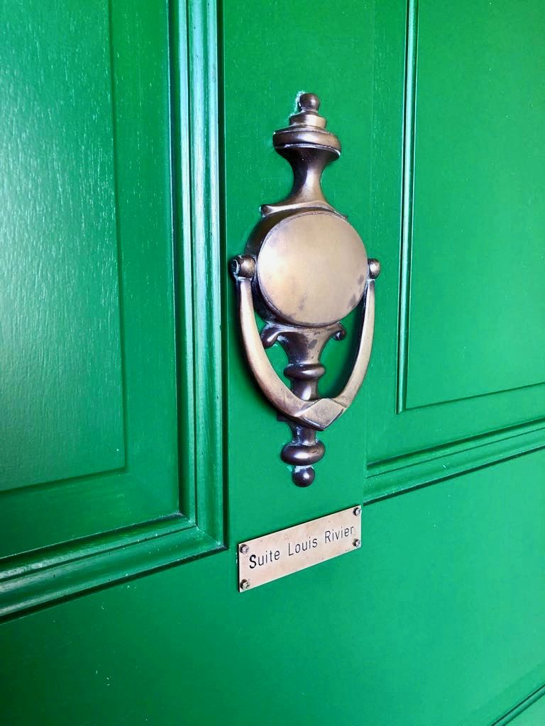 Une porte verte ornée d’un heurtoir en laiton et d’une plaque indiquant « Suite Louis Rivier ».