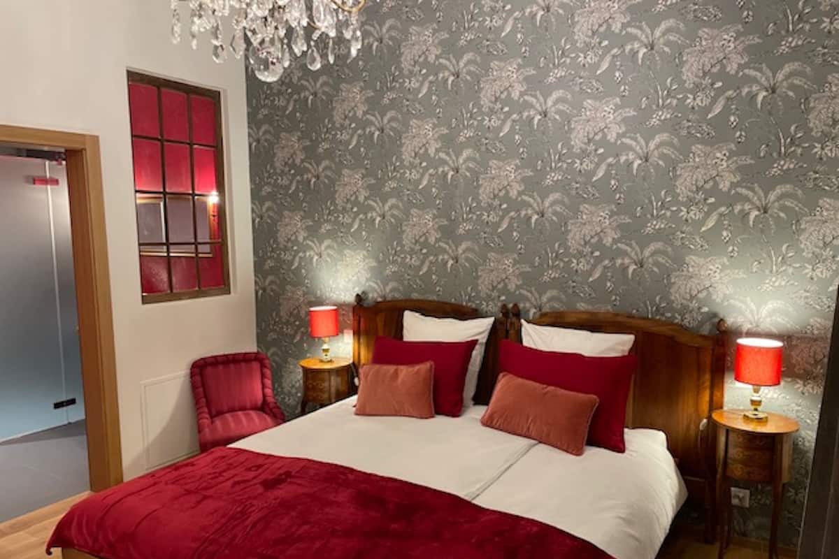 Ein Schlafzimmer mit einem großen Bett, zwei Nachttischen mit Lampen, einem Holz-Kopfteil, einem roten Sessel und einer Kronleuchter.