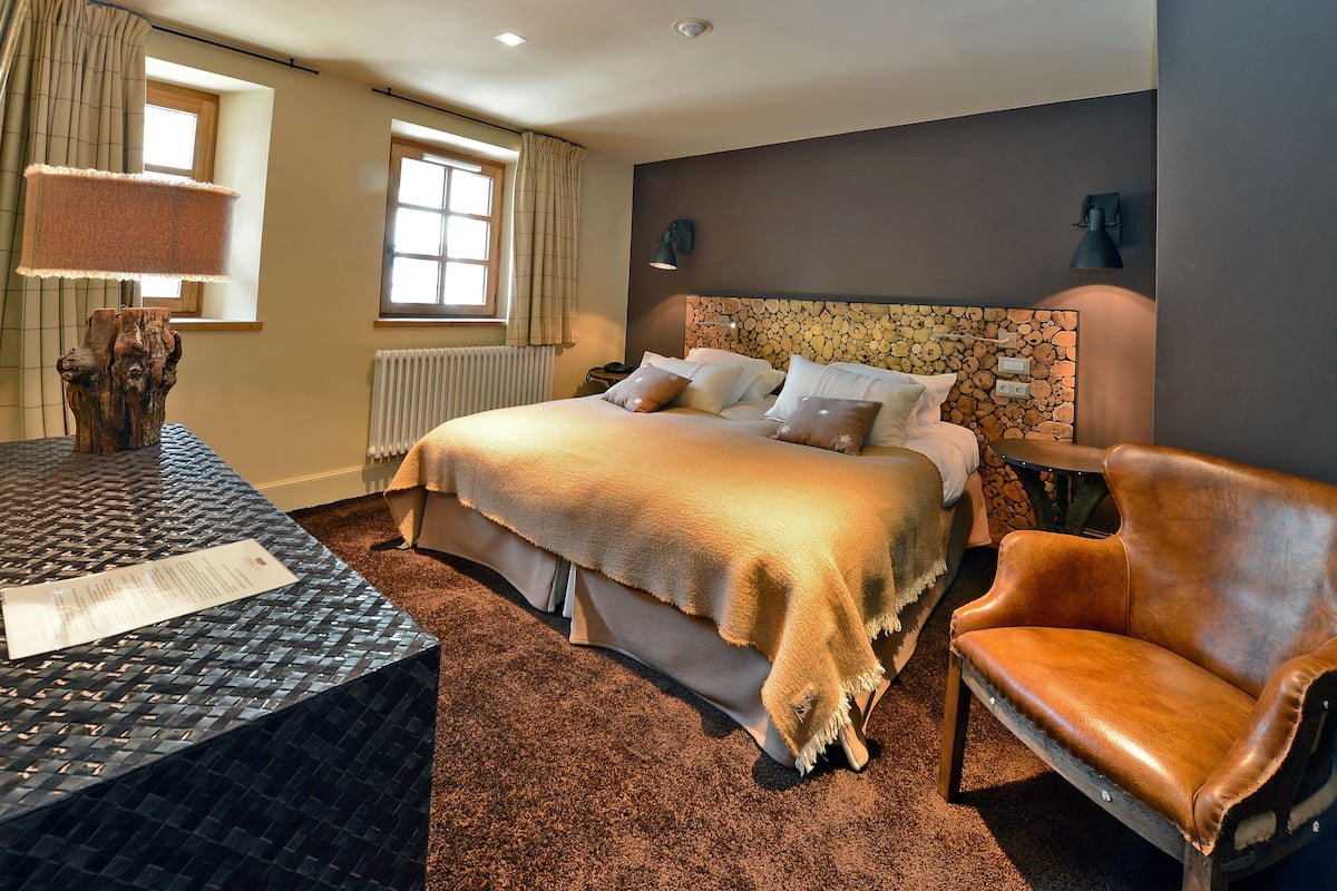 Une chambre d’hôtel avec un grand lit, un fauteuil en cuir, un bureau avec une lampe et une vue sur l’extérieur par une fenêtre.