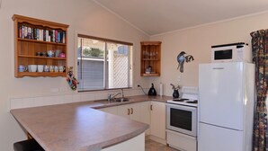 Eine kompakte Küche mit eingebautem Herd, einer Mikrowelle und einem Kühlschrank. Über der Spüle befinden sich Holzregale und ein Fenster mit Jalousien.