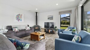 Ein modernes Wohnzimmer mit Sofa, Sesseln, Couchtisch, Fernseher, Kamin und einem großen Fenster mit Blick ins Freie.