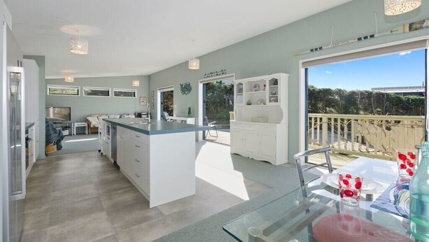 A spacious kitchen with a central island, a dining area with a glass table, and a balcony with a view of the trees. - The Blue Cottage with WiFi- Waipu Holiday Home (Waipu Cove)