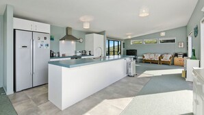 A modern kitchen with a central island, built-in appliances, and a dining area with a sofa and a TV. - The Blue Cottage with WiFi- Waipu Holiday Home (Waipu Cove)
