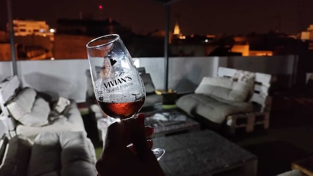 A hand holding a glass with "Vivian's Guest House" written on it, set against a backdrop of a modern outdoor seating area with sofas and a table. - Vivian's Guest House Auto Check-in (Jerez de la Frontera)