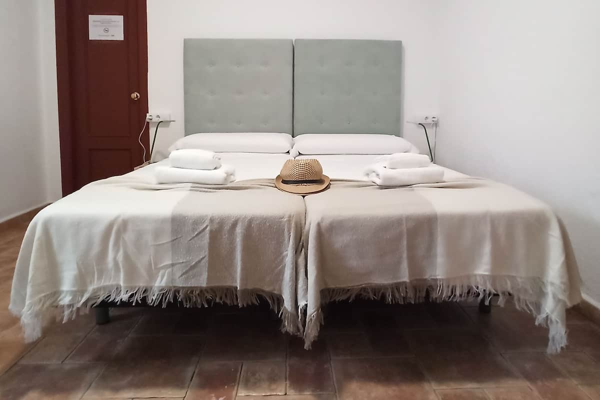 Una cama doble con cabecera gris, ropa de cama blanca y un pequeño sombrero sobre la cama.