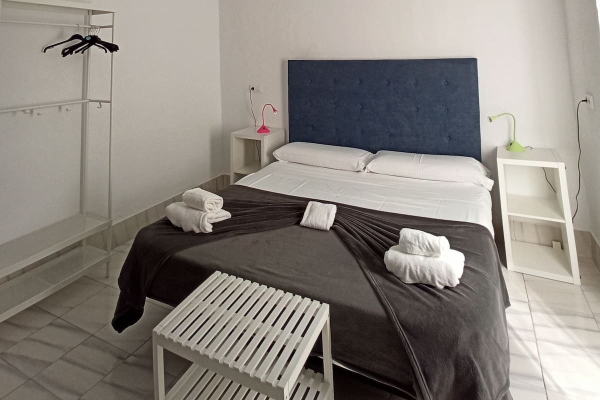 Habitación de hotel minimalista con una cama grande, una mesita de noche, una pequeña mesa y un perchero.