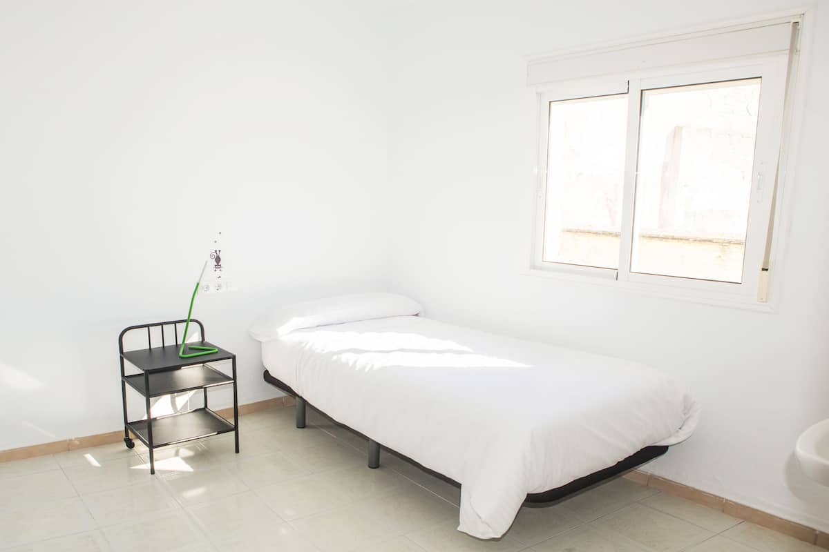 Una habitación pequeña y ordenada con una cama individual, una mesita con una planta y una ventana.