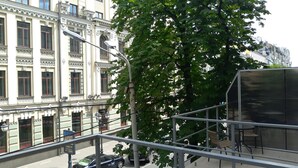 Vista de uma rua com um edifício de arquitetura clássica, uma árvore e uma varanda com mesas e cadeiras.
