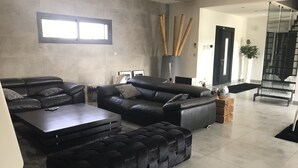 Una sala de estar moderna con un sofá de cuero negro, un reposapiés a juego y una mesa de centro. Hay una ventana grande con persianas, un jarrón alto con ramas de bambú y una escalera al fondo.