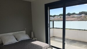 Un dormitorio con cama, mesita de noche, lámpara y vista a un cuerpo de agua y edificios a través de una puerta corrediza de vidrio.