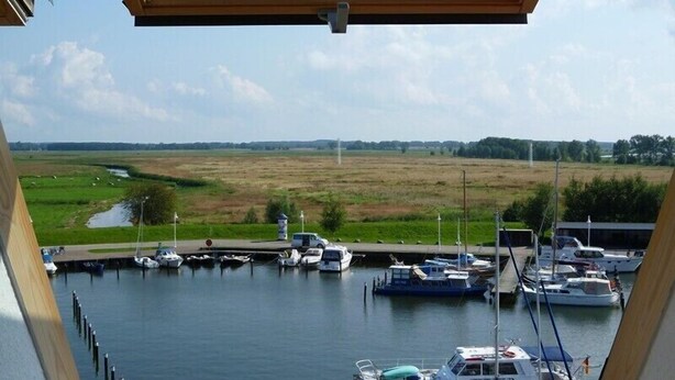 A view from a balcony overlooking a marina with boats docked, a grassy field, and a river in the background. - Wohnung Segelhafenidyll (Karlshagen)