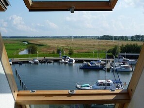 Blick vom Balkon auf einen Hafen mit vertäuten Booten, eine Wiese und ein Fluss im Hintergrund.