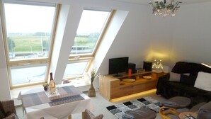 A modern living room with a dining table, wicker chairs, a sofa, and a TV. Large windows offer a view of the outdoors. - Wohnung Segelhafenidyll (Karlshagen)