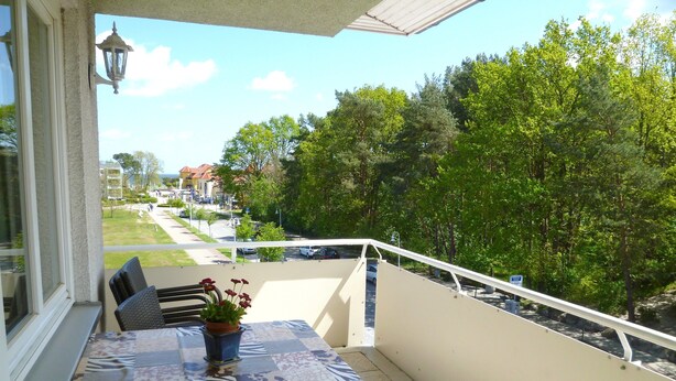 A balcony with a table and chairs, a potted plant, and a view of a residential street and trees. - Wohnung Ostseeblick (Karlshagen)