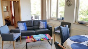 A living room with a black leather sofa, two blue armchairs, a glass coffee table, and a dining area with a table and chairs. - Wohnung Ostseeblick (Karlshagen)