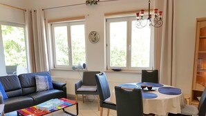 A living room with a sofa, chairs, a coffee table, and a dining table with placemats. There are windows with curtains, a chandelier, and a wall clock. - Wohnung Ostseeblick (Karlshagen)