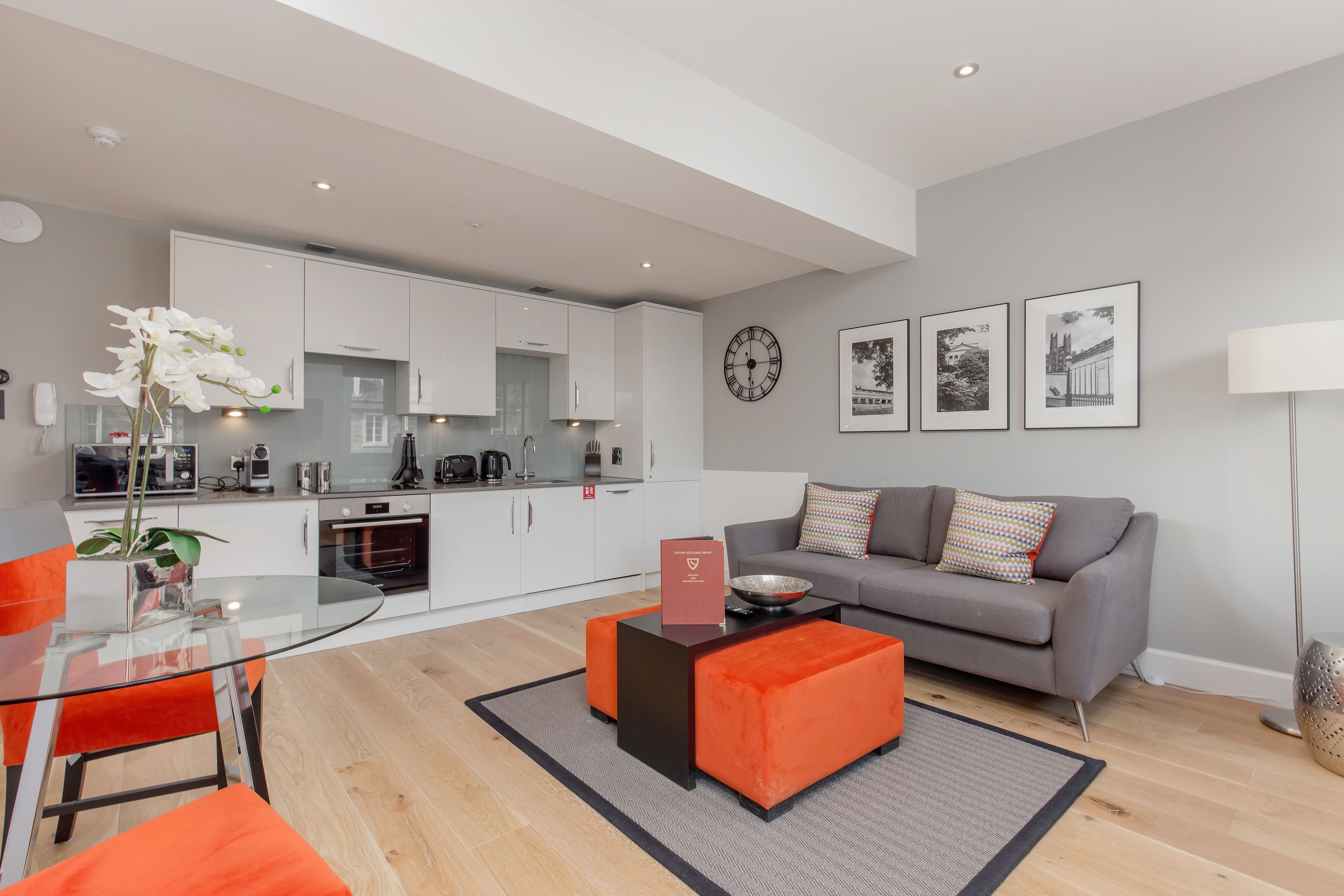 A modern living room with a glass dining table, orange ottomans, a grey sofa, and a kitchen in the background.