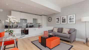 A modern living room with a glass dining table, orange ottomans, a grey sofa, and a kitchen in the background.