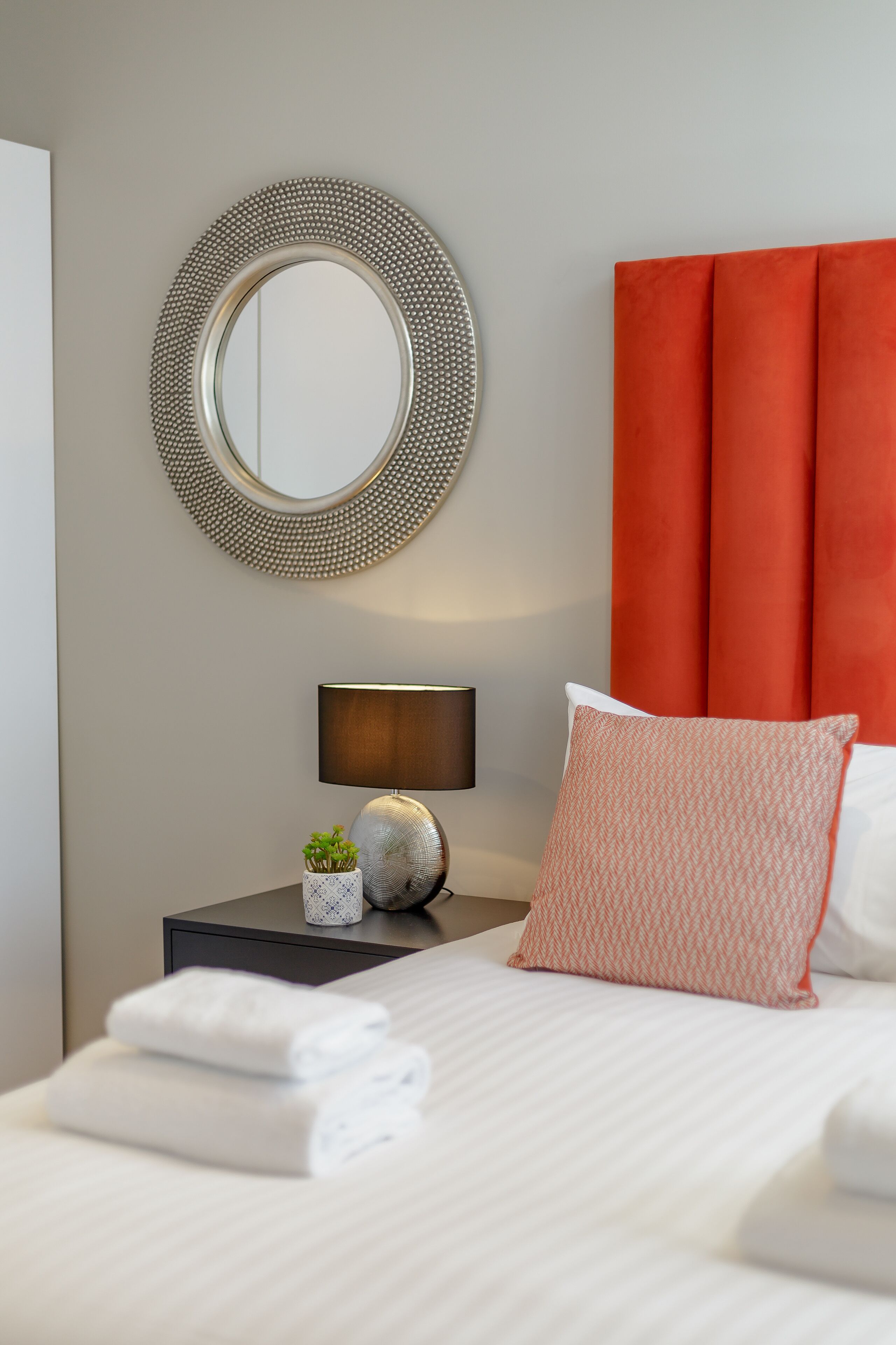 A neatly made bed with white linens, a round mirror with a beaded frame, a bedside lamp, and a red upholstered headboard.