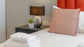 A neatly made bed with white linens, a round mirror with a beaded frame, a bedside lamp, and a red upholstered headboard.