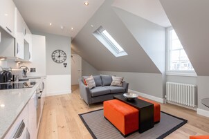A modern loft apartment with a kitchen, a gray sofa with patterned cushions, a black and orange ottoman, and a skylight.