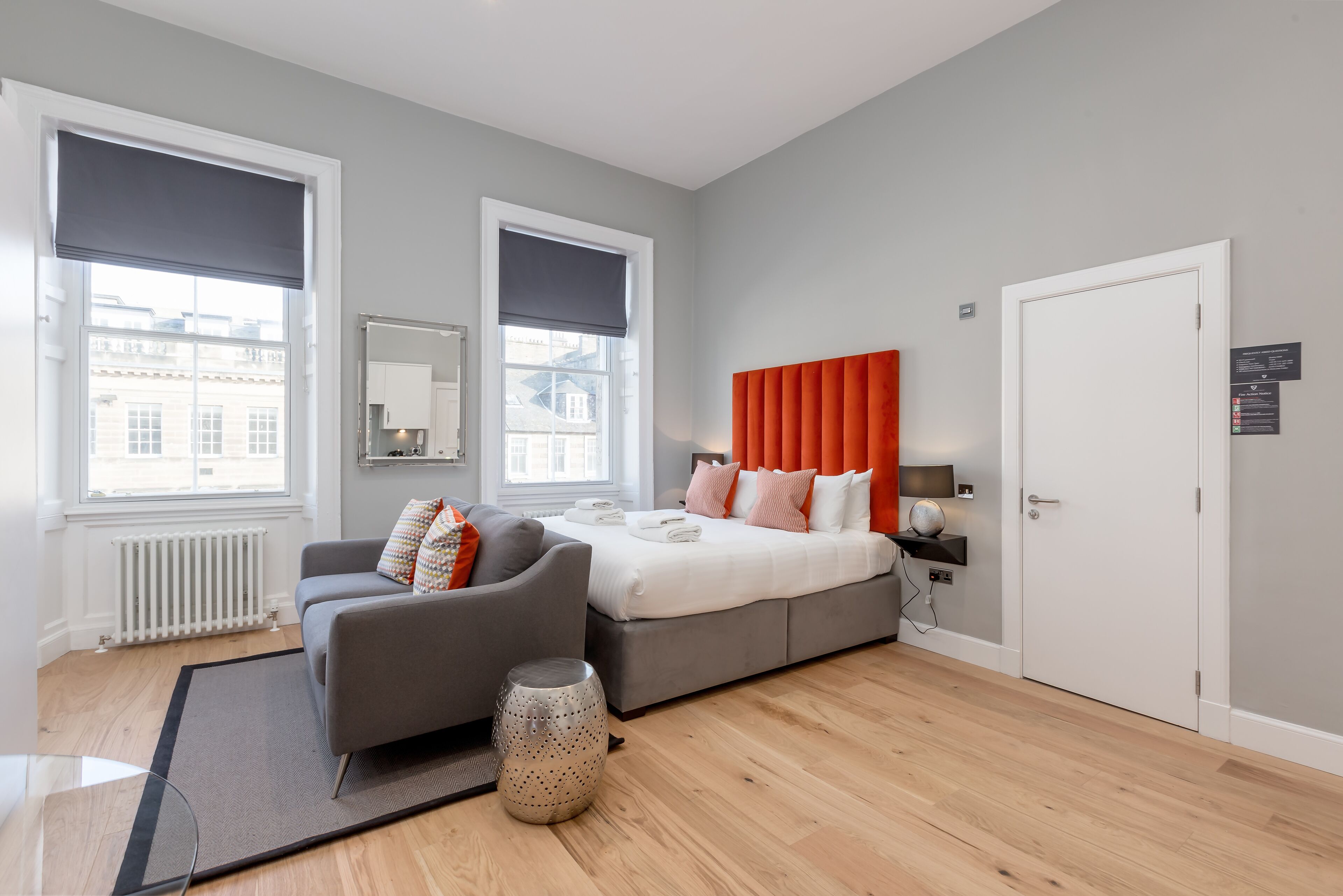 A modern bedroom with a grey sofa, a large bed with orange headboard, and a white door.