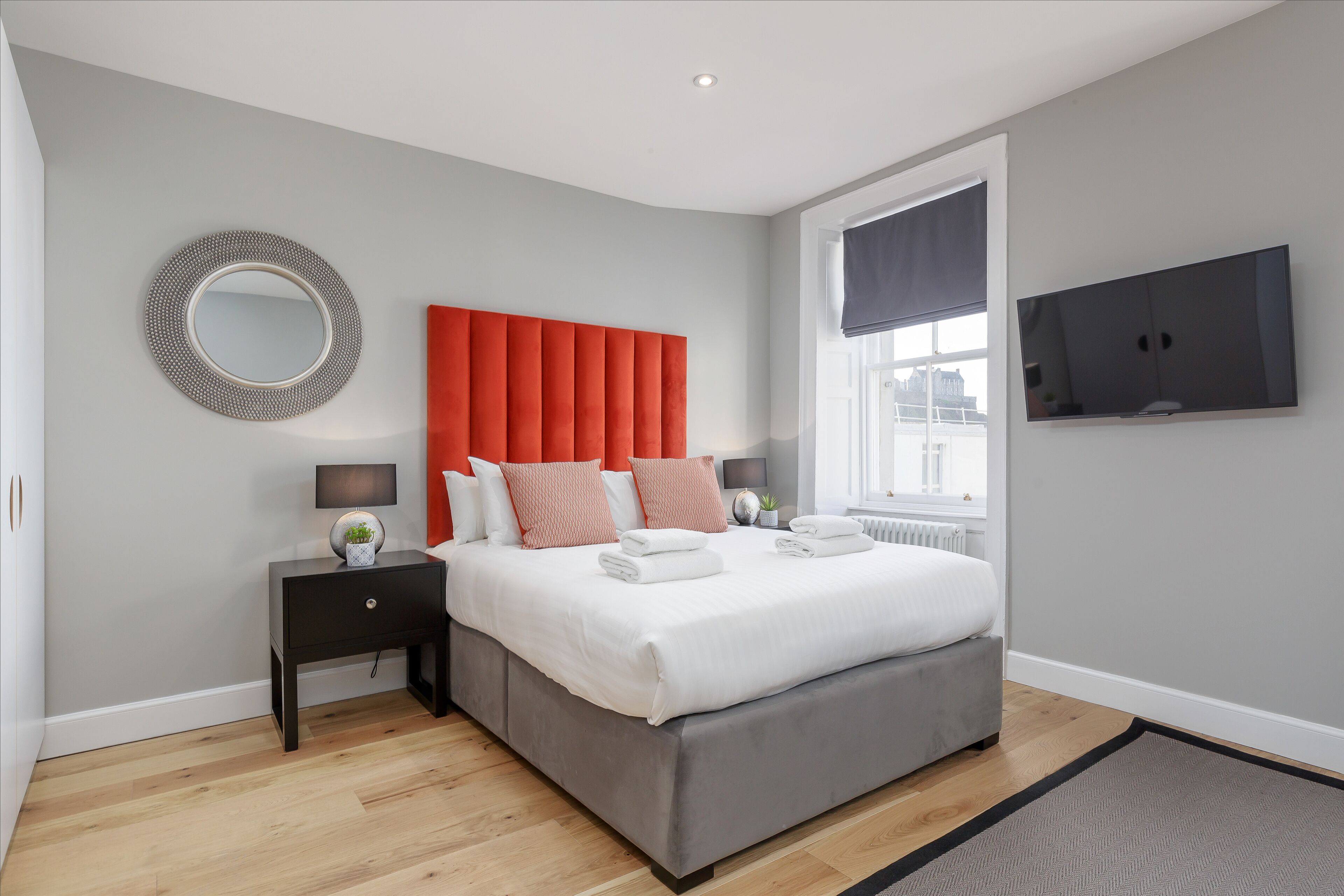 A modern bedroom with a grey upholstered bed, a red headboard, a round mirror, a flat-screen TV, and a window with a view of the city.