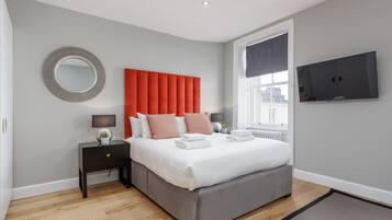 A modern bedroom with a grey upholstered bed, a red headboard, a round mirror, a flat-screen TV, and a window with a view of the city.