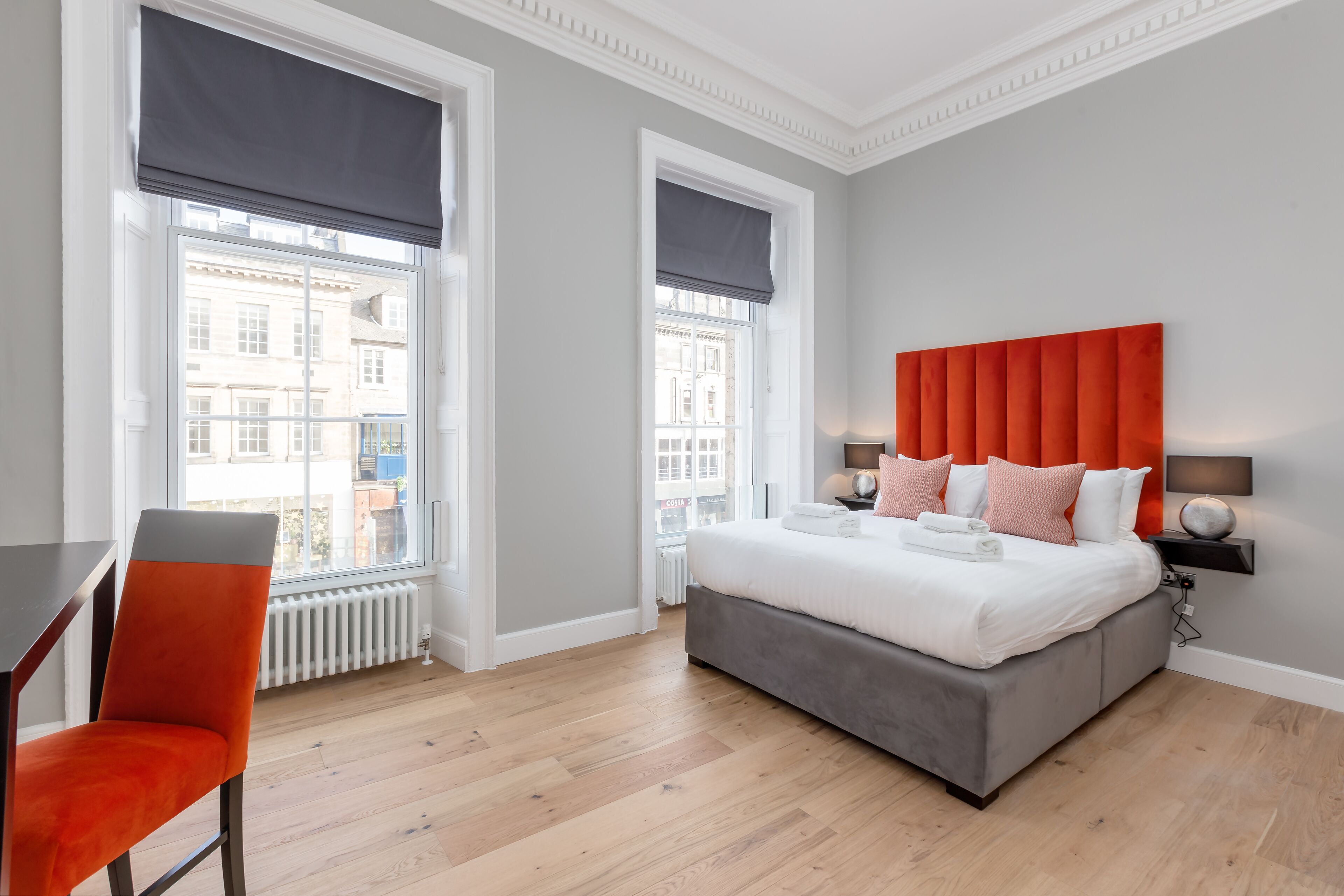 A modern bedroom with a large bed, a desk, and two windows with blinds.