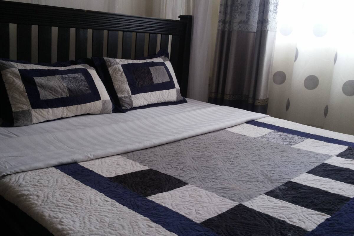 A bed with a quilted bedspread and two pillows, a wooden headboard, and a window with curtains.