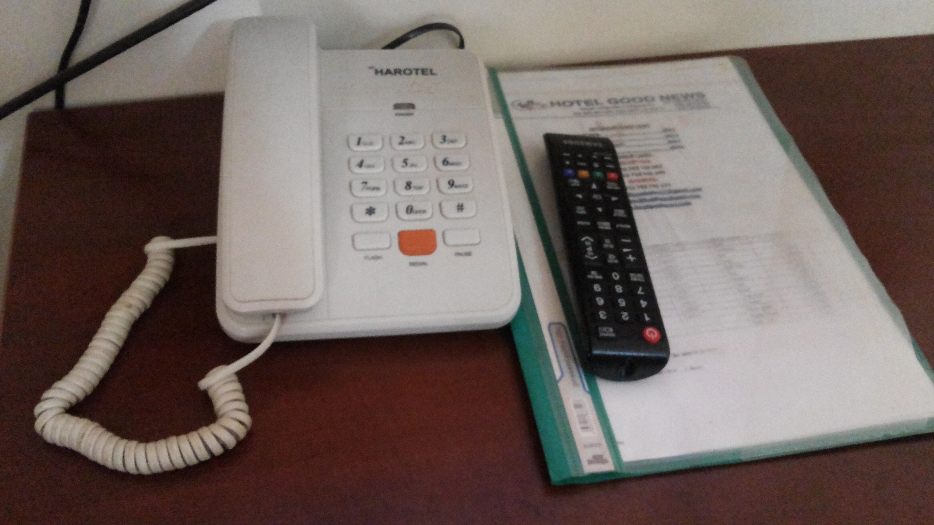 Un teléfono Harotel blanco y un control remoto sobre un escritorio con un folleto del hotel.