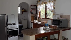 A kitchen with a stove, microwave, sink, and window with curtains. - Tropical Garden Self Catering (Mahé Island)